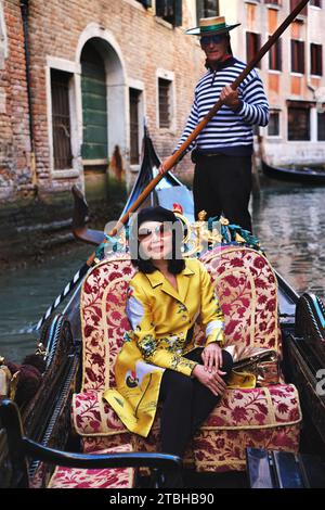 An stylish woman wearing a golden oriental coat sitting in a gondola being rowed by venetian gondolieri through picturesque canals of Venice, Italy Stock Photo