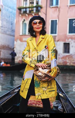An fashionable woman standing in a gondola wearing a golden Red-crowned crane coat riding through the picturesque canals - photoshoot, Venice, Italy Stock Photo