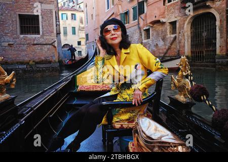 An elegant woman wearing a oriental coat in a gondola with golden horses being rowed past venetian architecture on the scenic canals of Venice, Italy Stock Photo