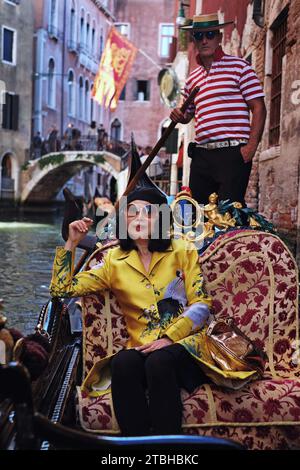 Asian woman wearing a golden coat sitting in a gondola being rowed by a red striped shirt wearing venetian gondolier on the canals of Venice, Italy Stock Photo