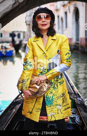 A glamourous  woman standing in a gondola wearing an oriental coat being rowed through picturesque canals past bridges & architecture of Venice, Italy Stock Photo