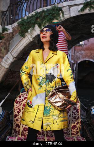 A stylish Asian woman standing in a gondola wearing a golden oriental coat  being rowed under a bridge by venetian gondolier Venice, Italy Stock Photo