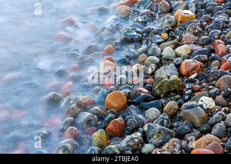 Blurred sea water of wave rolling over wet colourful pebbles on shingle beach / cobble beach Stock Photo