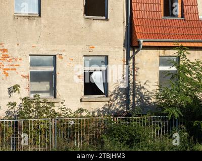 Abandoned building with broken windows and a damaged facade caused by vandalism. The house exterior is weathered and cracks are in the wall. Stock Photo