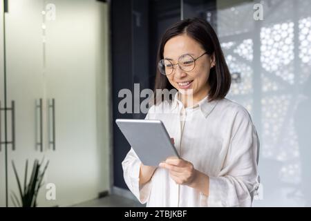 Young beautiful Asian woman with tablet computer standing near window, smiling businesswoman using online application, programmer testing new software. Stock Photo