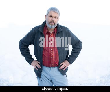 Man, 56 years, grey hair and beard, standing outside in winter with snow in background, Stock Photo