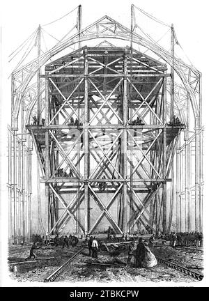 Progress of the Great Exhibition building: the traversing platform used in the construction of the nave, 1861. '...the most astonishing and the most extensive of [the] labour-saving contrivances is a gigantic travelling scaffold, which has been built on twelve wheels, to run on rails up and down the whole length of the centre nave. This huge structure is 60 ft. square and 100 ft. high, and weighs nearly 300 tons. Yet...four men with levers can move it with a certain amount of rapidity to any part of the works. It will be used in hoisting the upper columns, the huge circular wooden ribs of the Stock Photo
