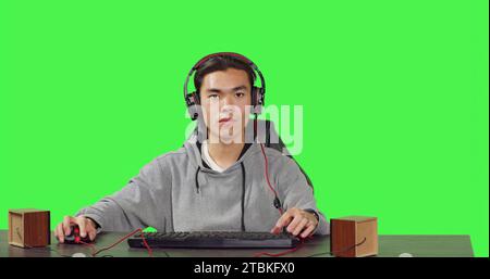 POV of asian man plays pc videogames at desk, sitting against greenscreen background. Gamer having fun with friends online on gaming contest, wearing headphones at workstation. Stock Photo