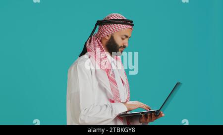 Person using laptop touchpad in studio, dressed in traditional muslim thobe and kufiyah. Middle eastern guy holding wireless computer and scrolling webpages or communicating on internet. Stock Photo