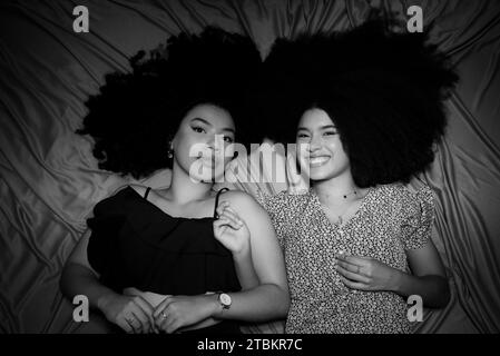 Half-closed black and white portrait of two women lying down with their curly hair and power spread on the floor cloth. Stock Photo