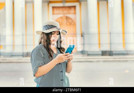 The girl wears a hat, stands in front of a Catholic church with a blurred background, is smiling and sending messages from her cell phone Stock Photo