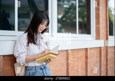 A pretty Asian female college student in casual clothes is taking notes in her book while standing outside of the building. University lifestyle conce Stock Photo