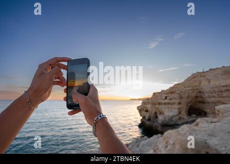 Golden Hour Moment: Young Man Hands Capturing the Ocean Sunset of beautiful cliff shore Stock Photo