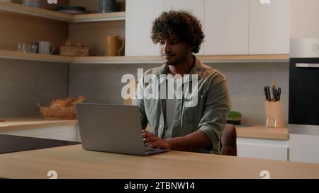 Concentrated Indian businessman typing computer working shopping online distant from home with laptop at kitchen serious focused freelancer Arabian Stock Photo