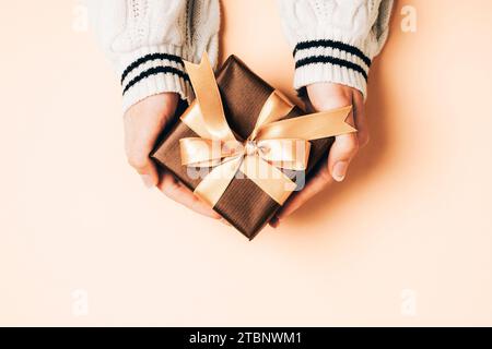 Girl's hands in white sweater holding brown gift box with golden bow on light peachy background. Christmas, Valentines day, Black friday sale concept. Stock Photo