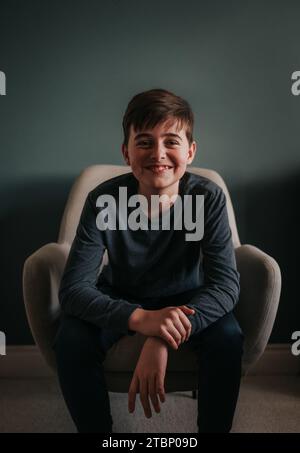 Portrait of happy boy sitting in chair indoors against blue wall. Stock Photo