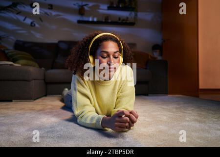 Young African American girl lying relaxed in living room listening to music wireless headphones.  Stock Photo