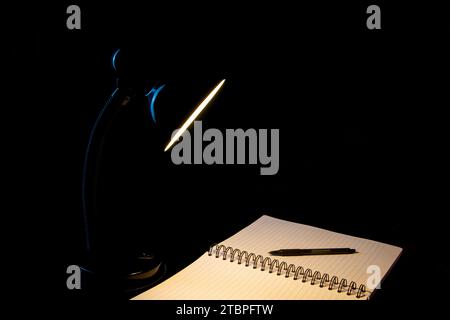 Closeup of a black desk lamp illuminating an open lined notepad and ballpoint pen on a black background Stock Photo
