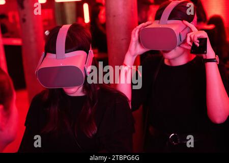 Two women in red-lit immersion, exploring virtual worlds together, amid the dynamic energy of a cutting-edge tech event Stock Photo