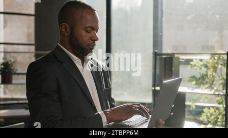 Focused mature adult office worker african american man middle-aged 40s ethnic businessman employee manager CEO specialist stand with laptop typing on Stock Photo