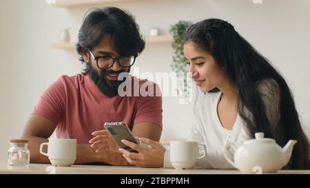 Diverse couple using smartphone arabian woman indian man in kitchen look mobile phone screen buying goods online order food delivery services internet Stock Photo