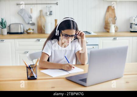Pre-teen 12s girl in headphones and eyeglasses sit at table e-learns, listen online course, audio lesson, get new knowledge, skills using internet and Stock Photo