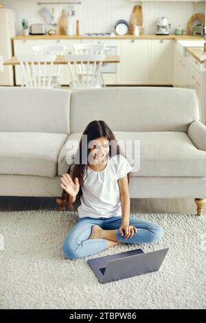 Attractive middle-eastern ethnicity pre-teen 12s girl sit on floor in living room wave hand greeting friend start videocall event use modern laptop. V Stock Photo