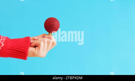 A woman's hand in a red New Year's sweater holds a Christmas ball, blue background. Christmas decor, red Christmas tree toy close-up. Banner, copy spa Stock Photo