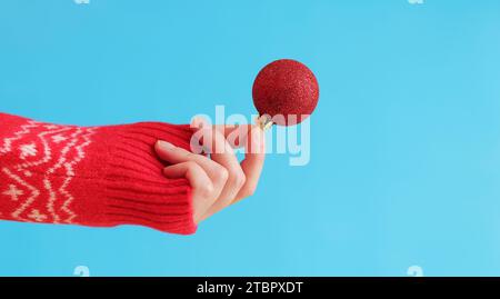 A woman's hand in a red New Year's sweater holds a Christmas ball, blue background. Christmas decor, red Christmas tree toy close-up. Banner, copy spa Stock Photo