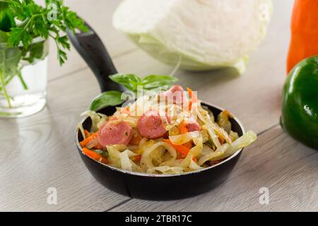 cooked fried cabbage with vegetables and sausages, in a frying pan on a wooden table. Stock Photo