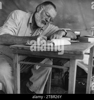 Photograph of Ernest Hemingway sitting at a table writing while at his campsite in Kenya circa 1953. Publisher Look Magazine. Stock Photo