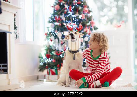 Christmas at home. Kids and dog under Xmas tree. Little boy and girl hug pet in Santa hat and open Christmas presents. Children play with animal. Stock Photo