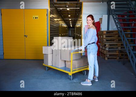 Red-haired woman stands with a cargo cart in a warehouse Stock Photo