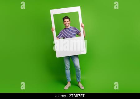 Full size photo of handsome good mood guy dressed denim pants striped shirt posing in photo frame isolated on green color background Stock Photo