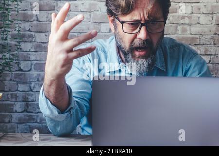 Small business freelance businessman shouting angry at the laptop in video call. One male adult people doing business online and managing problems. Exhausted worker in front of a computer shout angry Stock Photo