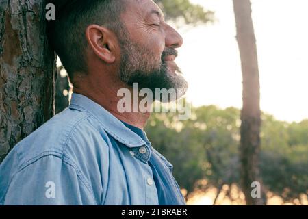 One adult man standing against a trunk tree having relaxation in outdoor leisure activity at the park alone. People and wellbeing connection with nature. Relax mental serene expression person mature Stock Photo