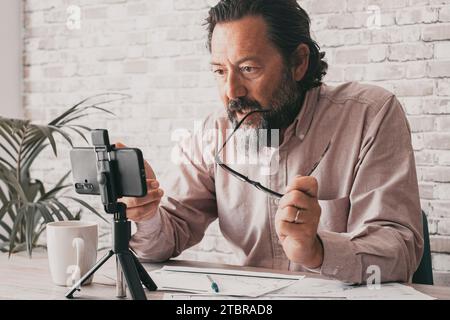 Small online business man talking in video call in front of a mobile phone on tripod in office home workplace. Paper documents on desk. Professional freelance working digital with cell communication Stock Photo