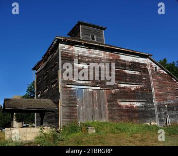 Water well sits in front of an unusual barn structure.  Weathered wood has faded from painted white to a dull gray.  Topped with a cupola, the Barn si Stock Photo