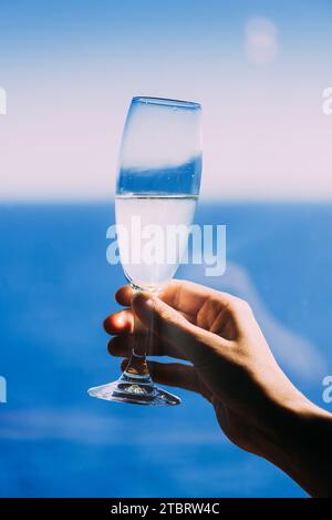 A young woman stands in a relaxed posture, her hand holding a glass of white wine as she looks out over a calm sea Stock Photo