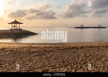 Sunrise over the sea, view from the sandy beach to the horizon, in the sea there are breakwaters with small temples, calm water with small waves and reflections on the tropical beach of Sanur, Bali, Indonesia Stock Photo