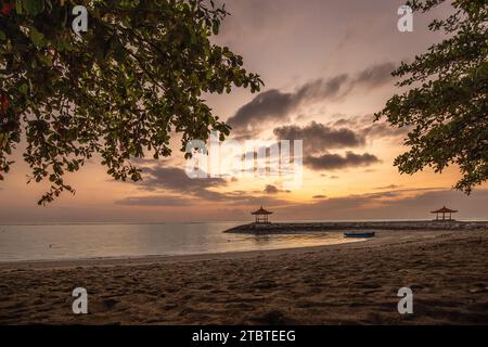 Sunrise over the sea, view from the sandy beach to the horizon, in the sea there are breakwaters with small temples, calm water with small waves and reflections on the tropical beach of Sanur, Bali, Indonesia Stock Photo