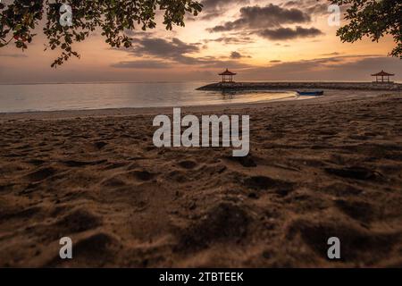 Sunrise over the sea, view from the sandy beach to the horizon, in the sea there are breakwaters with small temples, calm water with small waves and reflections on the tropical beach of Sanur, Bali, Indonesia Stock Photo