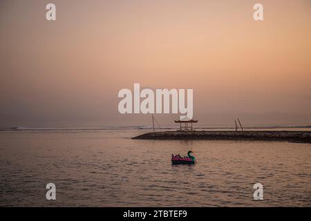 Sunrise over the sea, view from the sandy beach to the horizon, in the sea there are breakwaters with small temples, calm water with small waves and reflections on the tropical beach of Sanur, Bali, Indonesia Stock Photo