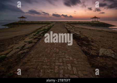 Sunrise over the sea, view from the sandy beach to the horizon, in the sea there are breakwaters with small temples, calm water with small waves and reflections on the tropical beach of Sanur, Bali, Indonesia Stock Photo