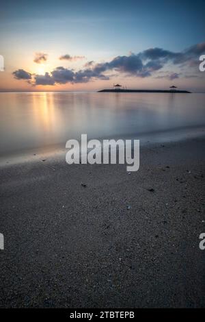 Sunrise over the sea, view from the sandy beach to the horizon, in the sea there are breakwaters with small temples, calm water with small waves and reflections on the tropical beach of Sanur, Bali, Indonesia Stock Photo