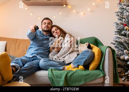Heterosexual young couple with their lapdog sitting on couch at home and watching TV during Christmas holidays. Stock Photo