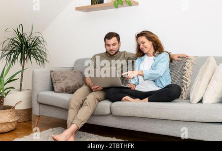 Young couple string together on couch at home and using laptop. Online shopping, e-commerce. Stock Photo