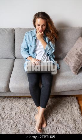 High angle view of young brunette woman sits on couch at home and using her laptop. Stock Photo