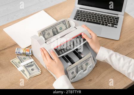 Woman putting dollar banknotes into cash counting machine at table in office, closeup Stock Photo