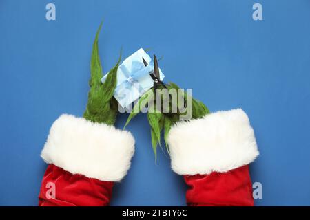 Green hairy hands of creature in Santa costume with gift box and scissors on blue background Stock Photo